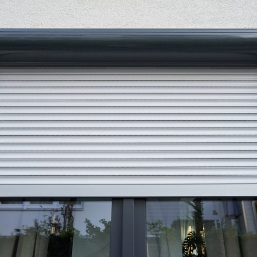 Do roller shutters block out noise? A close-up image of a grey window roller shutter in a half-closed position, demonstrating the potential for noise reduction in a residential setting.